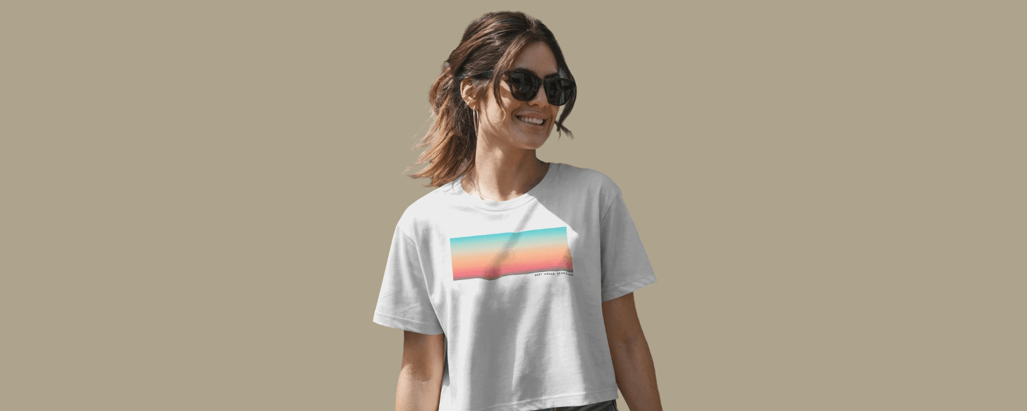 Woman in sunglasses wearing a white Best Human Beaming t-shirt with a pastel ombre rectangle design, smiling against a taupe background. Showcases sustainable, empowering fashion.