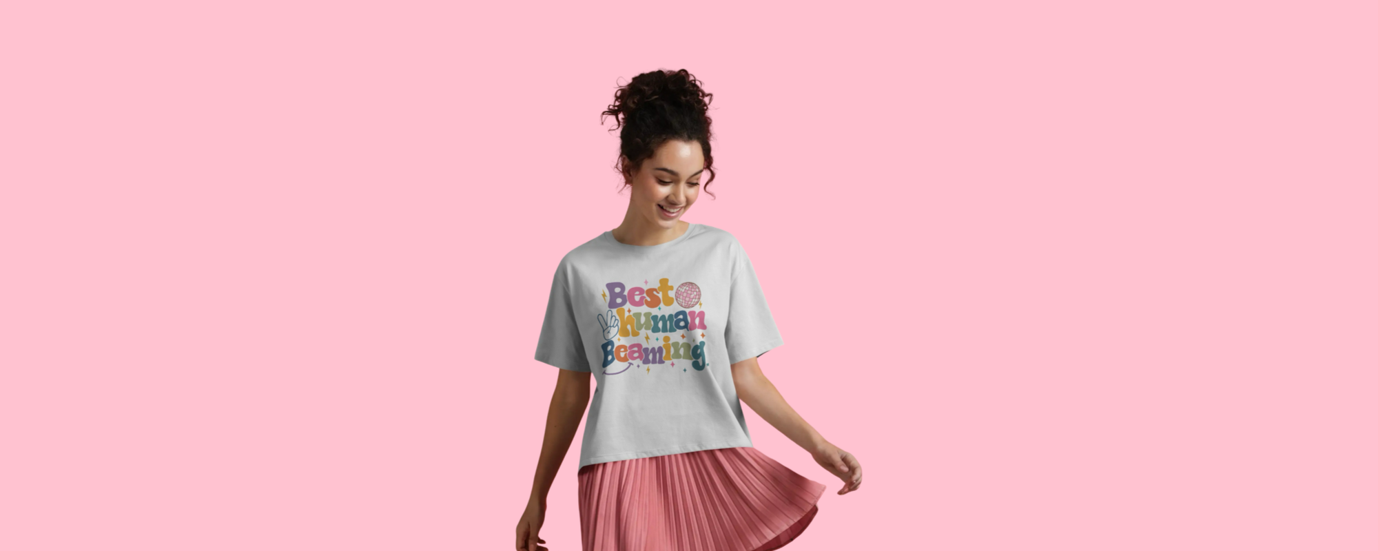 Woman in a white Best Human Beaming t-shirt with a colourful retro logo and pleated pink skirt, smiling and moving playfully against a pink background. Celebrates joyful, inclusive style.
