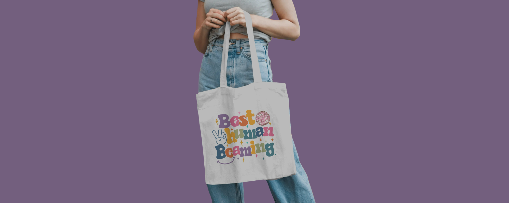 Person holding a white Best Human Beaming tote bag with colourful retro logo, peace sign, disco ball, and stars. Fun, eco-friendly accessory for self-expression.