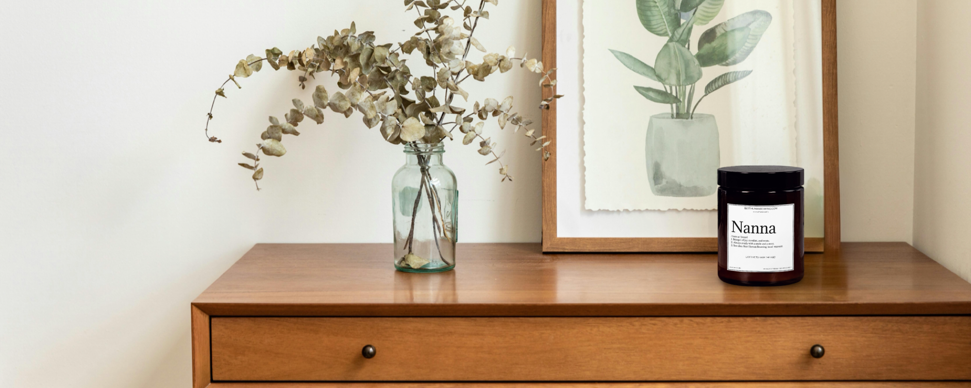 Glass jar with dried eucalyptus branches on a wooden table beside a framed botanical print. Minimalist, calming home decor style reflecting eco-conscious and natural living.