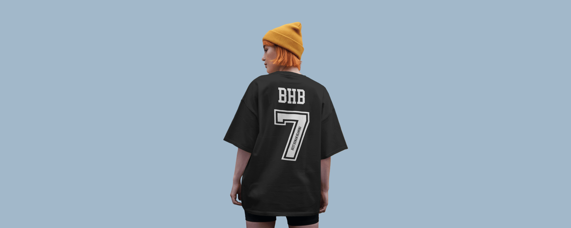 Person with orange hair and yellow beanie wearing a black Best Human Beaming t-shirt with bold “BHB” and large number 7, seen from behind. Modern, sporty, and inclusive style.