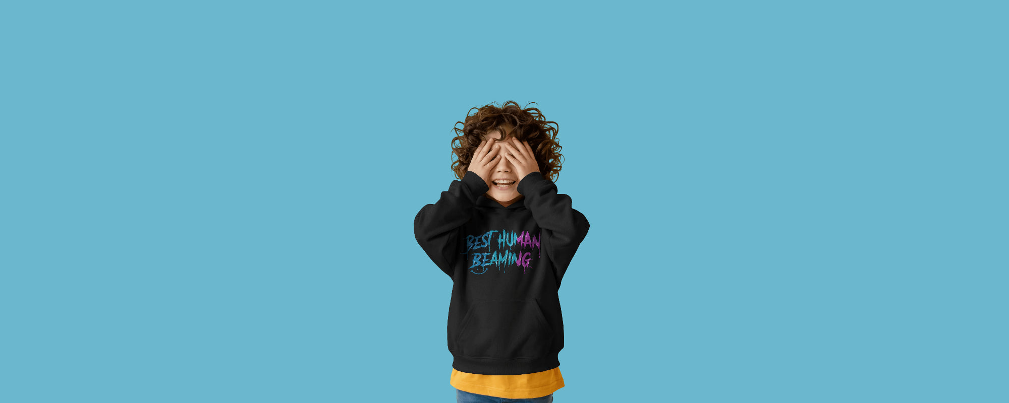 Smiling child in a black Best Human Beaming hoodie with blue and pink graffiti logo, playfully covering their eyes, on a blue background. Fun, empowering, and inclusive kids’ fashion.