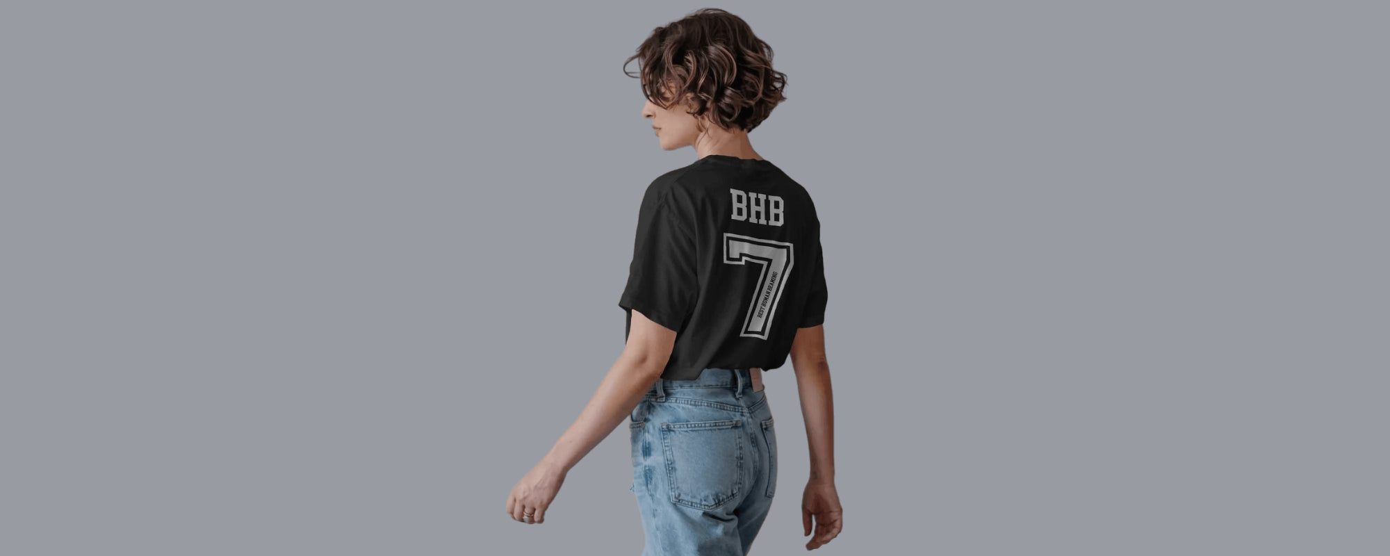 Woman in a black Best Human Beaming t-shirt, back view, with bold white “BHB” and a large number 7. Modern, sporty design highlights confidence and sustainable fashion.