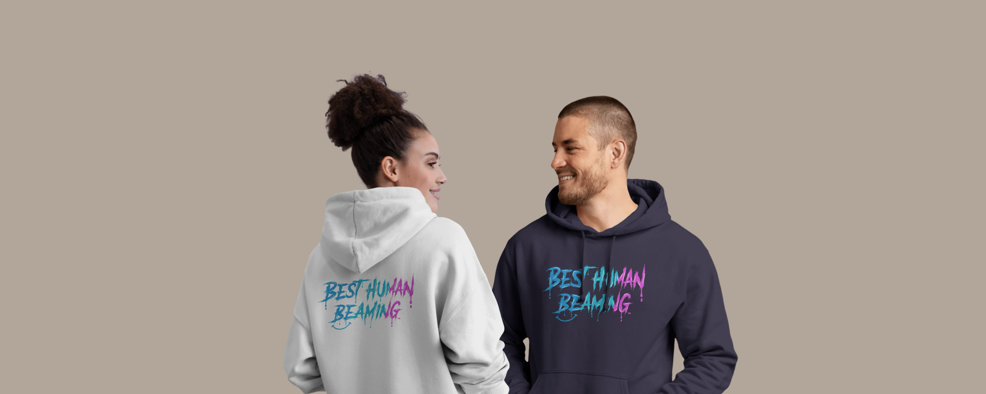 Two people in Best Human Beaming hoodies with blue and pink graffiti logo, one in white and one in navy. Smiling at each other, representing joyful, unisex, and empowering style.