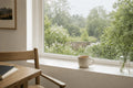 Cream ceramic mug of tea on a bright windowsill overlooking a lush garden in gentle rain, with soft natural light, wooden chair and calm Scandinavian-inspired interior.