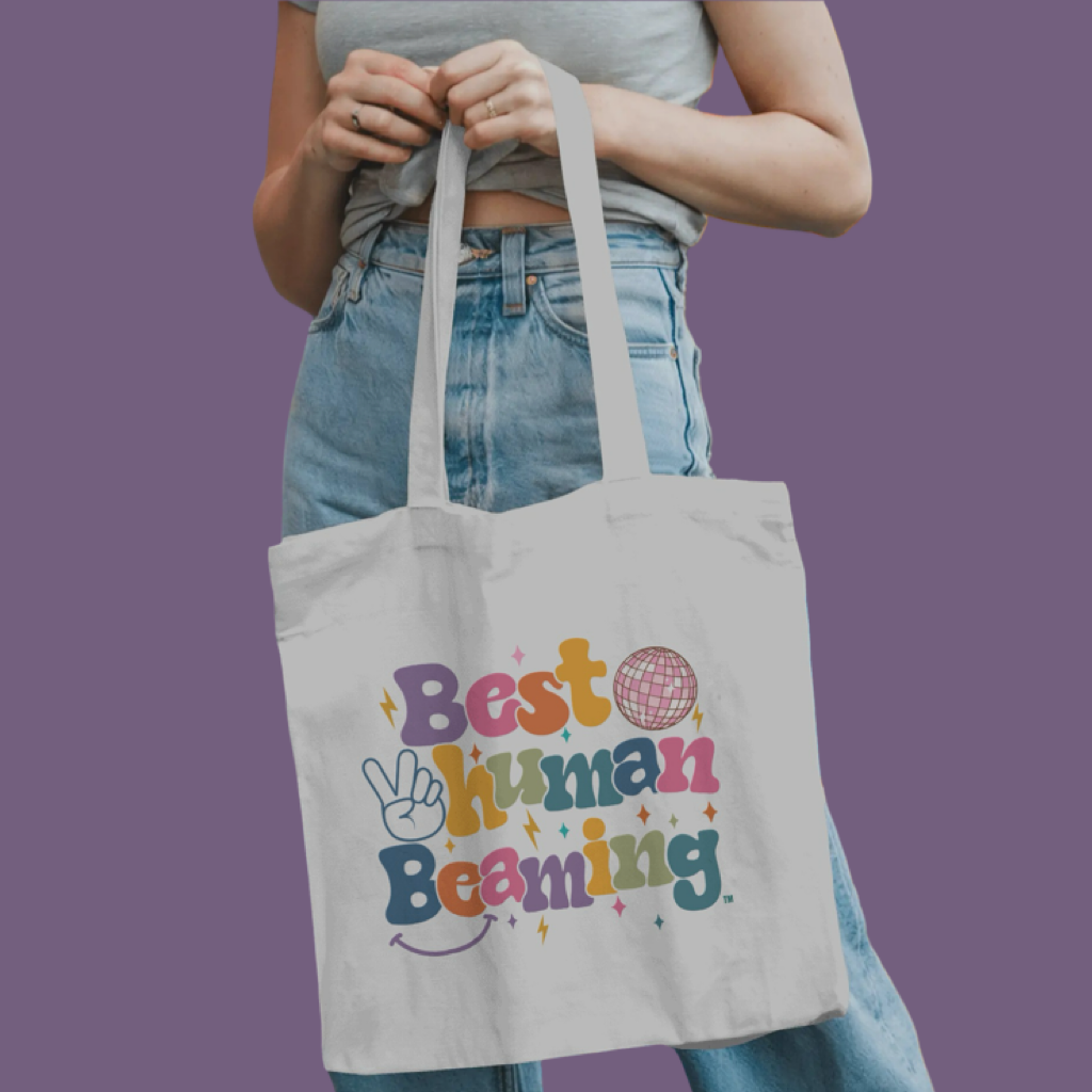 Person holding a white Best Human Beaming tote bag with colourful retro logo, peace sign, disco ball, and stars. Fun, eco-friendly accessory for self-expression.
