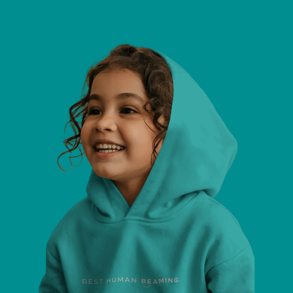Smiling young girl in a teal Best Human Beaming hoodie with minimalist logo. The look is joyful and inclusive, reflecting confidence and uniqueness.