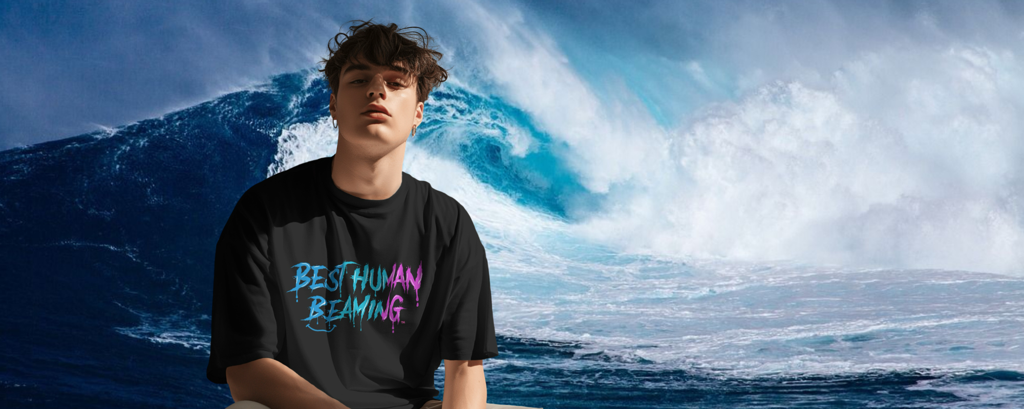 Young man in a black Best Human Beaming t-shirt with blue and pink graffiti logo, standing in front of a powerful ocean wave. Bold, eco-friendly streetwear for confident self-expression.