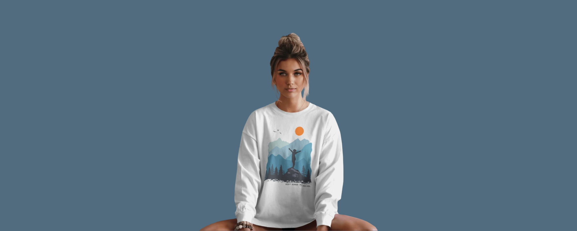 Woman sitting cross-legged in a white Best Human Beaming sweatshirt with a mountain and sun graphic. Design celebrates adventure, empowerment, and self-worth in sustainable fashion.